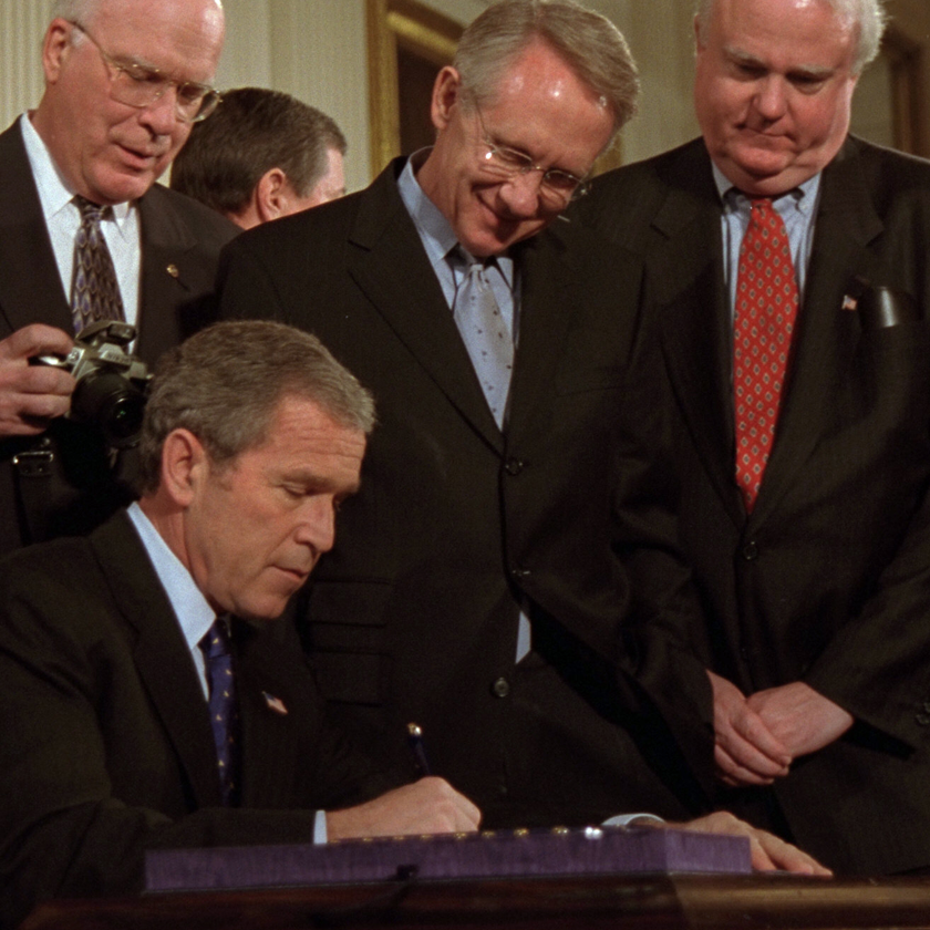 President Bush signing with a pen at a desk while surrounded by white men in suits looking on.
