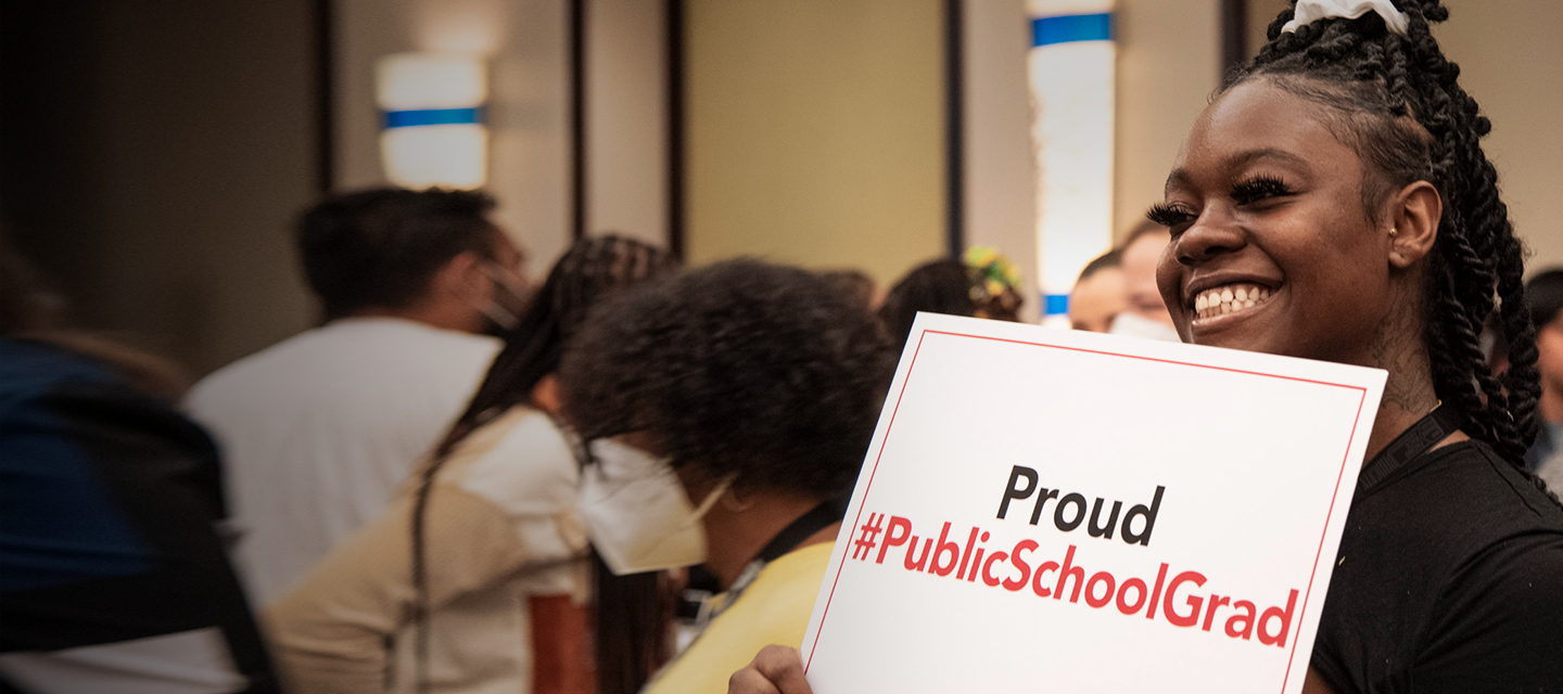 Young adult holding a sign that reads "Proud (hashtag) Public School Grad"