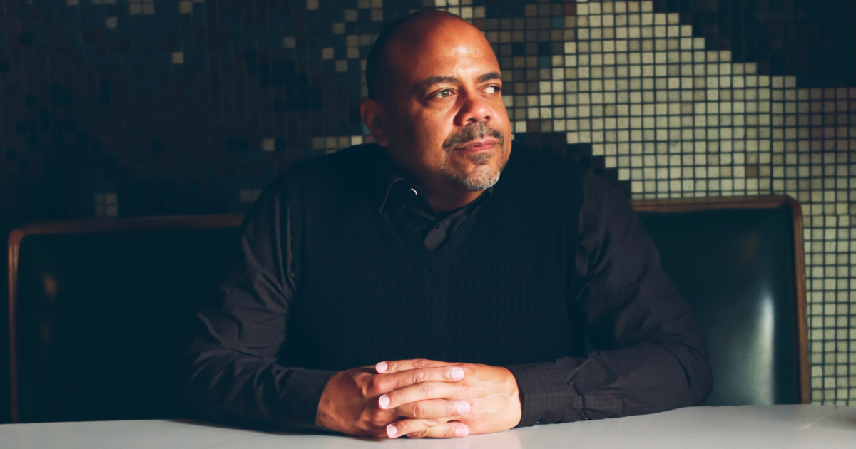 Race Forward Executive Vice President Eric Ward poses with hands crossed on a desk, wearing a black shirt against a black and olive tiled background.