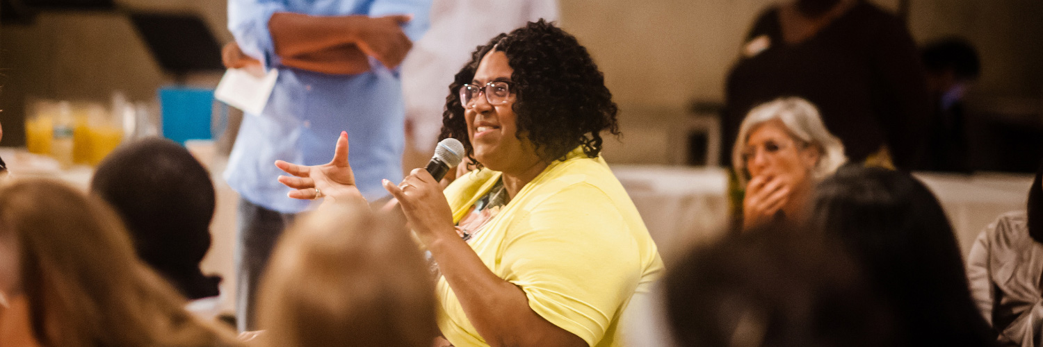 Seated person speaking into mic with other individuals looking on.