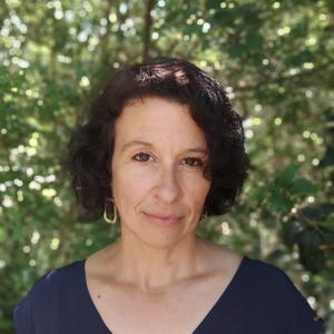 Photo of a white woman with wavy dark brown chin-length hair and brown eyes, wearing hoop earrings and a dark blue shirt outside against a blurred background of sun-dappled green bushes.