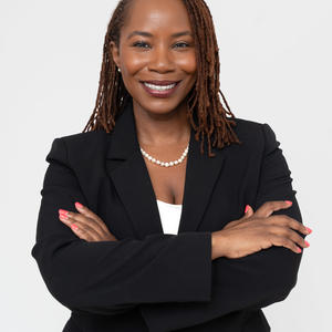 Photo of Ritha Pierre, a Black woman with medium chestnut-brown loc’d hair and a radiant smile. Her high cheekbones and confident, professional presence make her very approachable and poised.