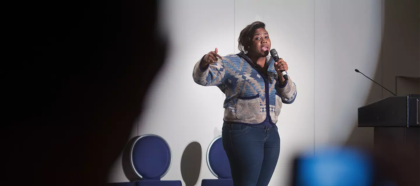 Young Adult speaking into a microphone on stage in front of a seated crowd